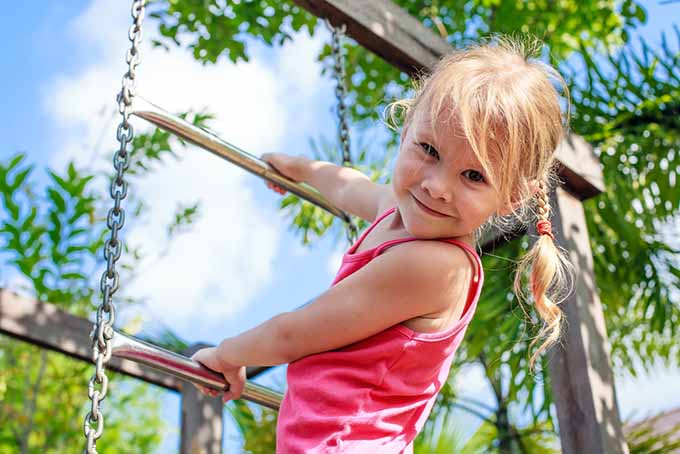 夏日，一个小女孩微笑着爬上游乐场的金属梯子，背景是蓝天和云彩。