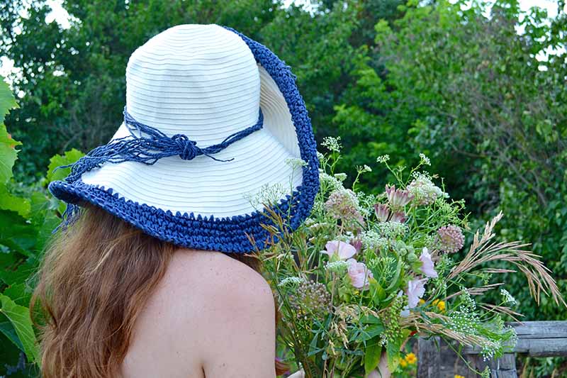 一名年轻女子裸露肩膀，戴着蓝白相间的帽子，手里拿着一束新摘的花，背景是树木。