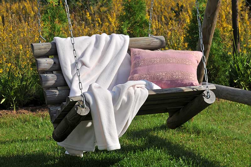 这是一张在阳光明媚的花园里拍摄的，带有粉红色靠垫和米色靠垫的乡村木制悬挂长椅的特写。