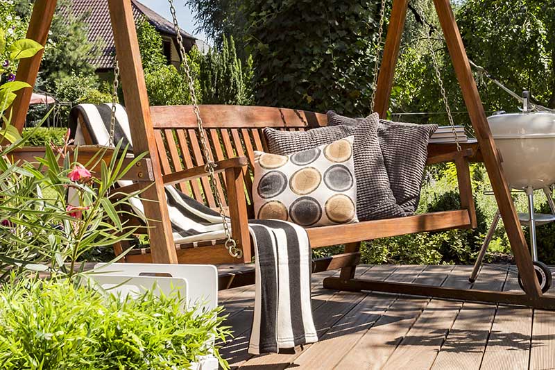 一个近距离的木制长椅座椅，散落的靠垫和条纹地毯，设置在一个以花园为背景的木制甲板上。
