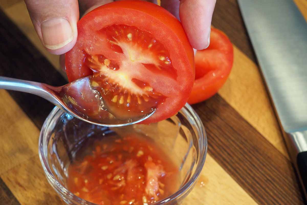 密切的横向图像框架的一只手从上拿着番茄已被削减了一半,和一个勺子挖出种子。