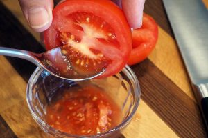一个近距离的水平图像，从框架的顶部，一只手拿着一个被切成两半的西红柿，一个勺子舀出种子。背景是一个木制表面和一把金属刀。